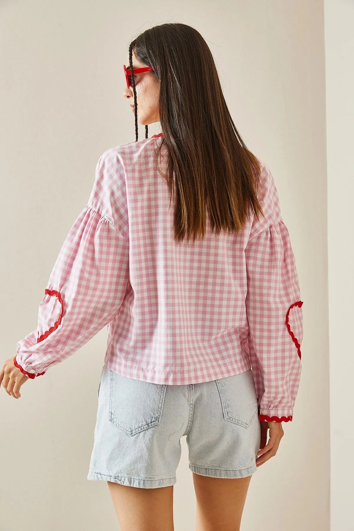 Pink - Women's pink and white gingham tie-front blouse featuring long sleeves, contrast red scalloped trim, and playful heart-shaped appliqués, styled with light wash denim shorts.