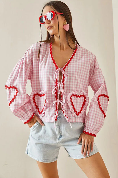 Pink - Women's pink and white gingham tie-front blouse featuring long sleeves, contrast red scalloped trim, and playful heart-shaped appliqués, styled with light wash denim shorts.
