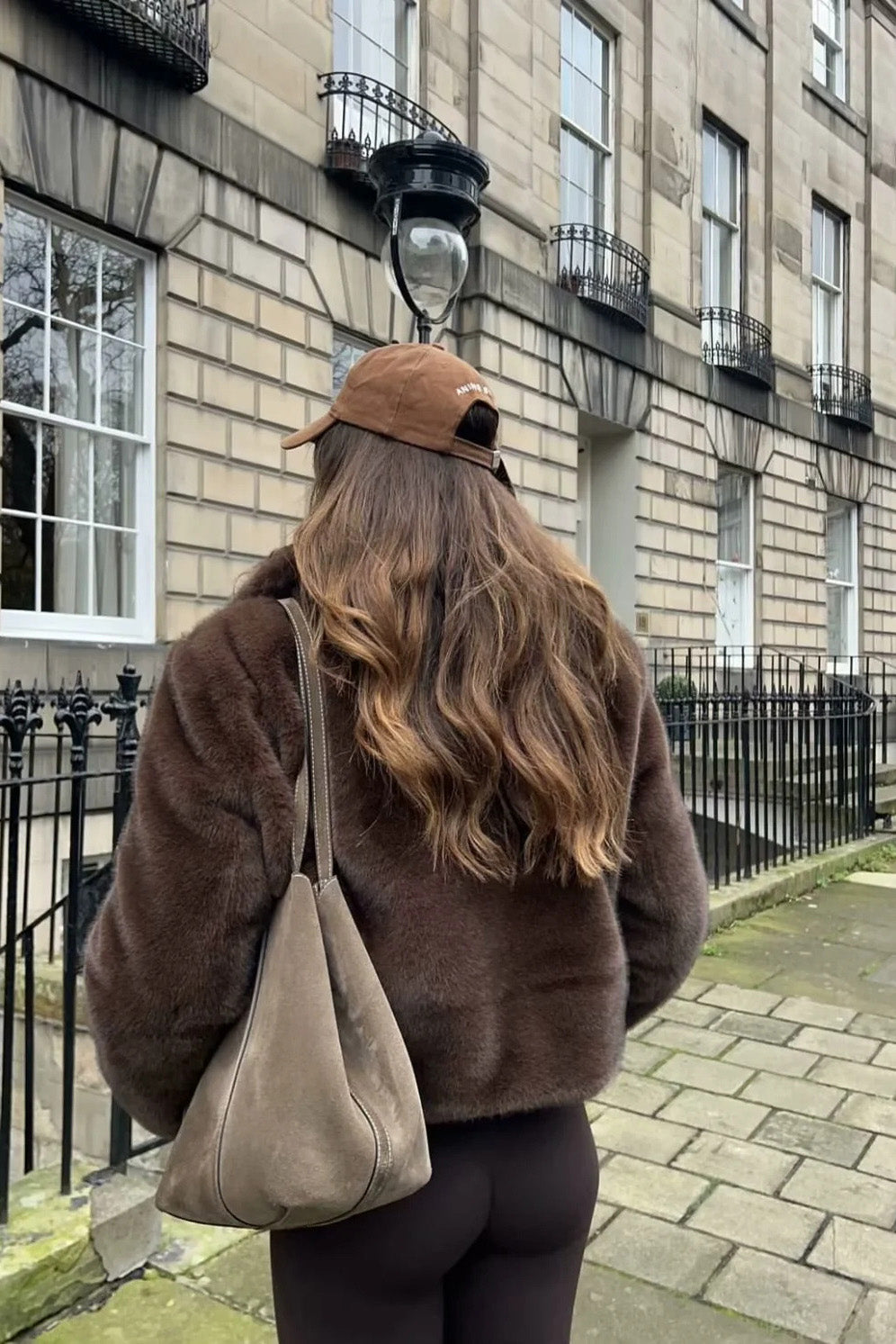A woman standing outdoors on stone steps wearing a dark brown, plush faux fur cropped jacket with a wide collar, styled with a mini skirt and tall black boots.