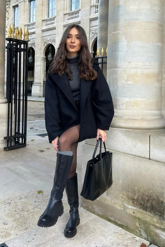 Black - A woman wearing a black oversized short coat with drop shoulders and a notched lapel, styled over a grey turtleneck and black shorts for a chic city look.