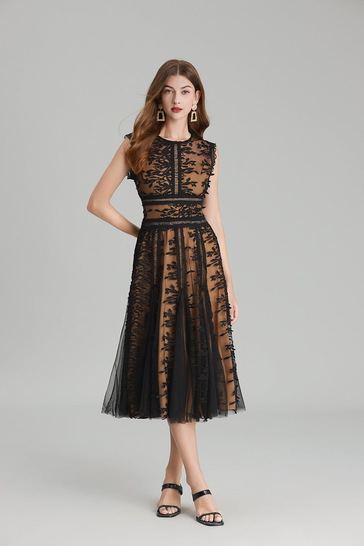 Full-length view of a woman wearing a sleeveless, sheer midi dress in pink and nude, featuring elaborate pink botanical embroidery in vertical panels and a flared tulle skirt.