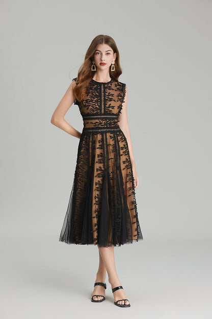 Full-length view of a woman wearing a sleeveless, sheer midi dress in pink and nude, featuring elaborate pink botanical embroidery in vertical panels and a flared tulle skirt.