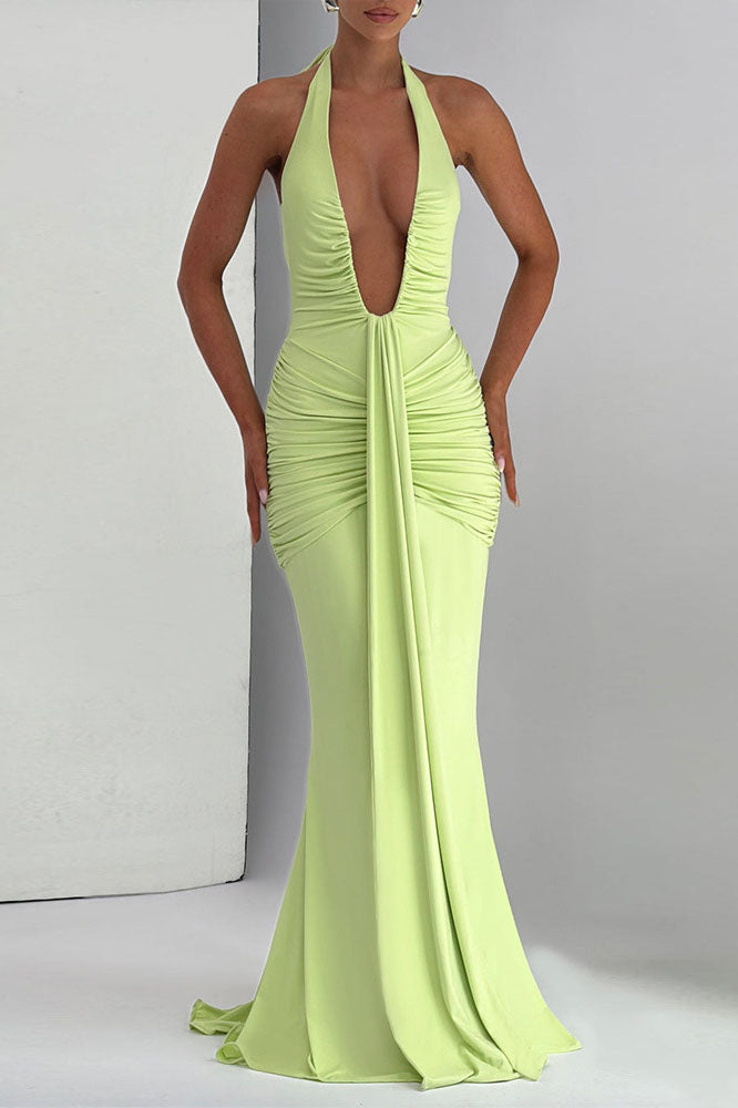 Green - A woman models a form-fitting, deep red halterneck maxi dress with a plunging neckline and extensive ruching, standing elegantly in a formal garden setting.
