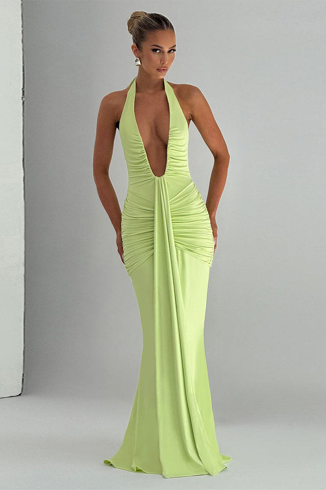 Green - A woman models a form-fitting, deep red halterneck maxi dress with a plunging neckline and extensive ruching, standing elegantly in a formal garden setting.