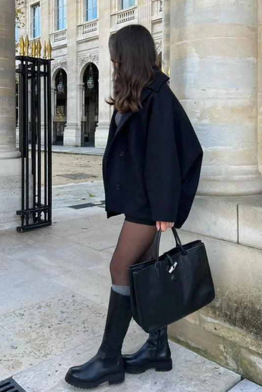 Black - A woman wearing a black oversized short coat with drop shoulders and a notched lapel, styled over a grey turtleneck and black shorts for a chic city look.