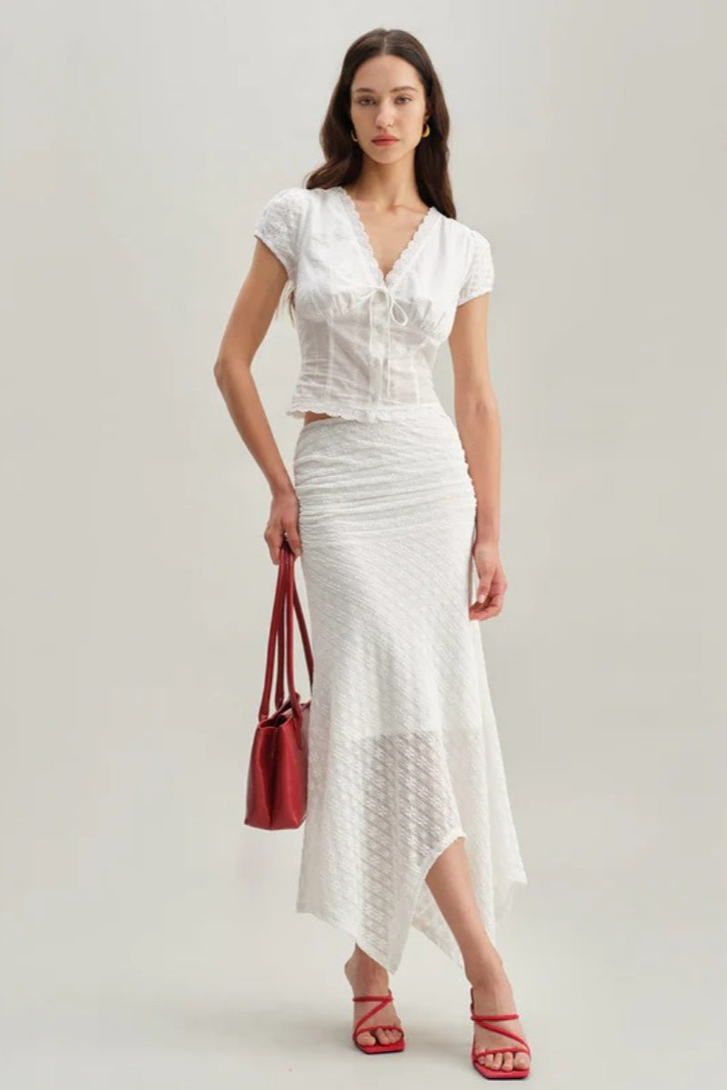 White - A woman wearing a white, short-sleeved corset-style blouse featuring a deep V-neckline with scalloped lace trim, a tie-front detail, and subtle tonal embroidery.