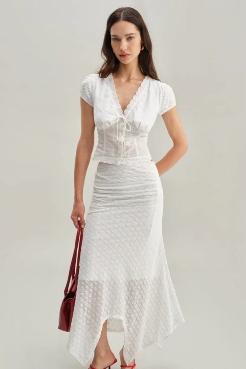 White - A woman wearing a white, short-sleeved corset-style blouse featuring a deep V-neckline with scalloped lace trim, a tie-front detail, and subtle tonal embroidery.