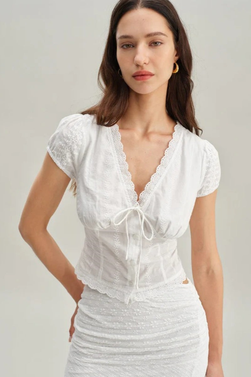 White - A woman wearing a white, short-sleeved corset-style blouse featuring a deep V-neckline with scalloped lace trim, a tie-front detail, and subtle tonal embroidery.