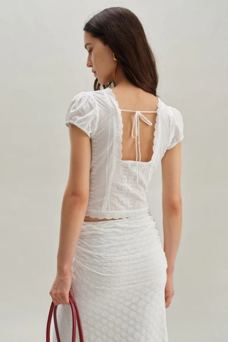 White - A woman wearing a white, short-sleeved corset-style blouse featuring a deep V-neckline with scalloped lace trim, a tie-front detail, and subtle tonal embroidery.