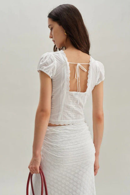 White - A woman wearing a white, short-sleeved corset-style blouse featuring a deep V-neckline with scalloped lace trim, a tie-front detail, and subtle tonal embroidery.