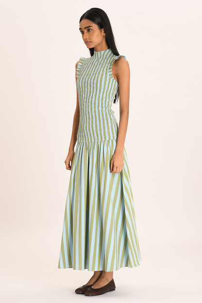A woman models a sleeveless maxi dress with a high neckline and a fully smocked bodice. The dress features a pattern of vertical light blue and olive green stripes, ruffled shoulder details, and a full A-line skirt.