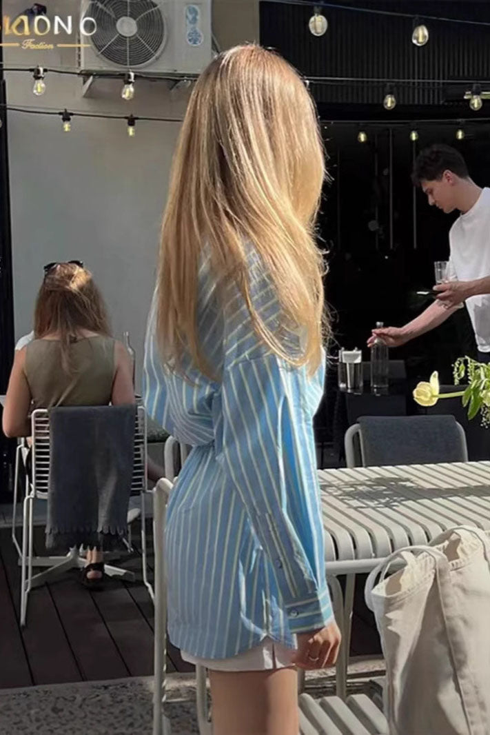 A woman wearing a blue and pale yellow vertically striped mini shirt dress featuring a collared neckline, button-down front, long sleeves, and a thick elasticated waistband.