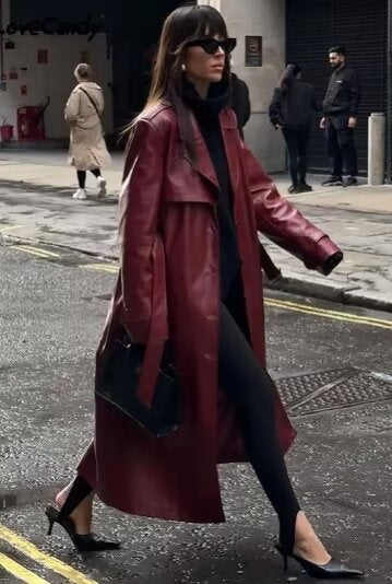 A woman walking down a city street wearing a long, deep burgundy faux leather trench coat with a tie belt, styled over a black turtleneck and black leggings.