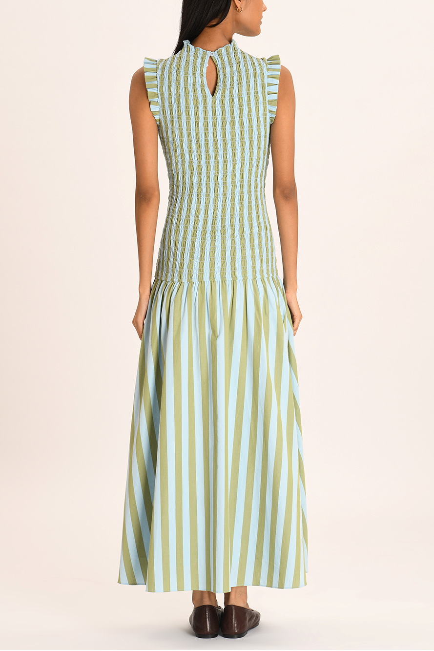 A woman models a sleeveless maxi dress with a high neckline and a fully smocked bodice. The dress features a pattern of vertical light blue and olive green stripes, ruffled shoulder details, and a full A-line skirt.