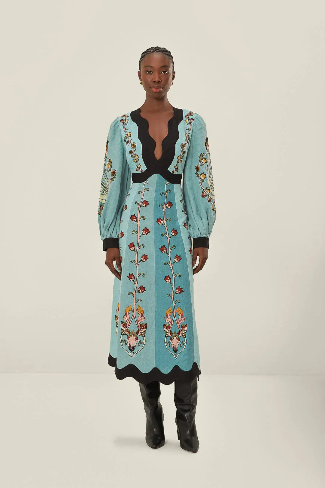 Full-length studio shot of a woman wearing a light blue, long-sleeved midi dress with a vibrant botanical print. The dress features a deep V-neckline, bishop sleeves, and contrasting black scalloped trim at the neck, waist, cuffs, and hem.