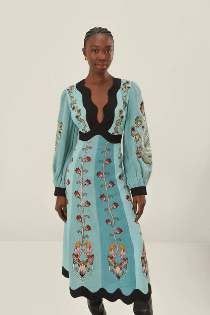 Full-length studio shot of a woman wearing a light blue, long-sleeved midi dress with a vibrant botanical print. The dress features a deep V-neckline, bishop sleeves, and contrasting black scalloped trim at the neck, waist, cuffs, and hem.
