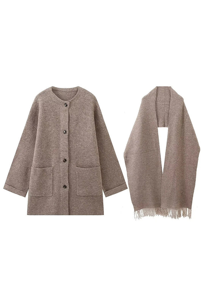 Grey - A woman wearing a thick, taupe oversized longline winter coat featuring a built-in scarf neckline, dark button closures, and large patch pockets.