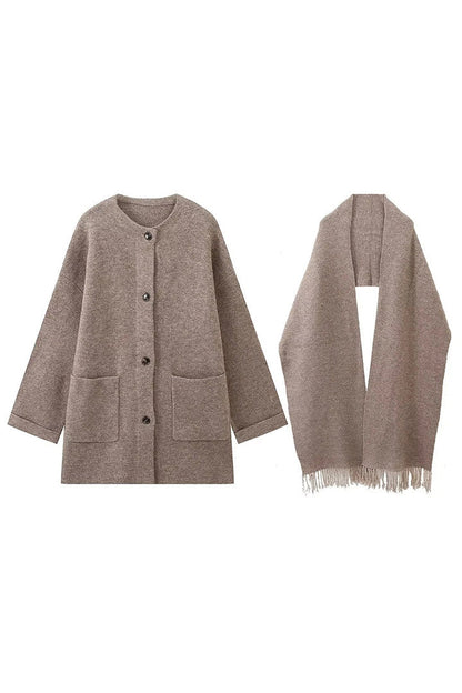 Grey - A woman wearing a thick, taupe oversized longline winter coat featuring a built-in scarf neckline, dark button closures, and large patch pockets.