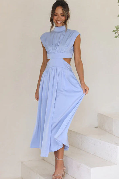 A woman walks outdoors in a pale yellow midi dress, its full skirt flowing around her. The dress features a high funnel neck, cap sleeves, and cutouts at the waist.