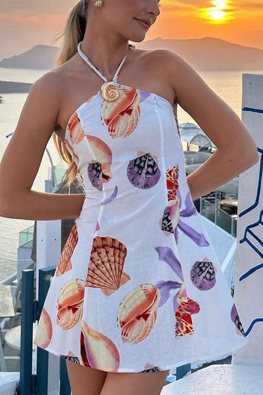 A woman wearing a white, A-line halter mini dress featuring a colorful all-over seashell print. The braided halter strap is secured at the front with a large, gold-tone spiral shell embellishment. She is pictured from the waist up on a balcony overlooking the ocean at sunset.