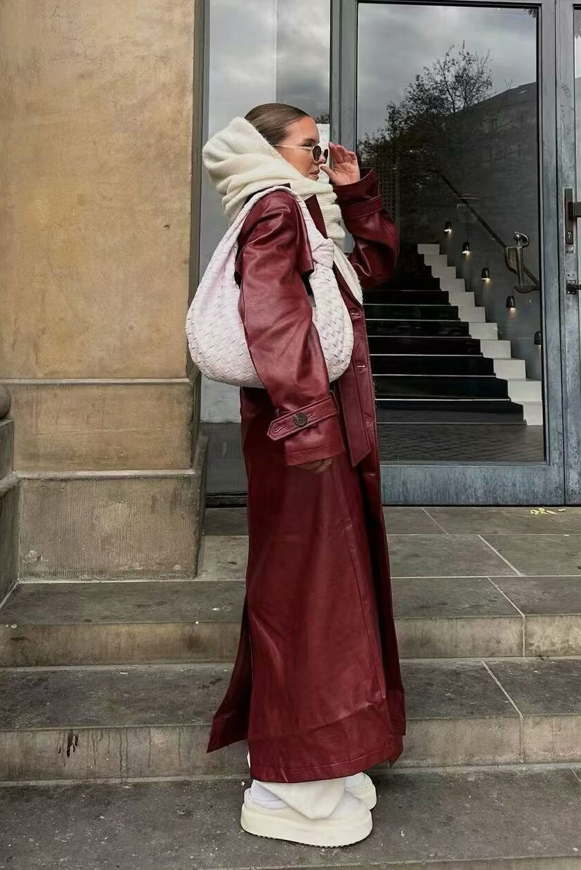 A woman walking down a city street wearing a long, deep burgundy faux leather trench coat with a tie belt, styled over a black turtleneck and black leggings.