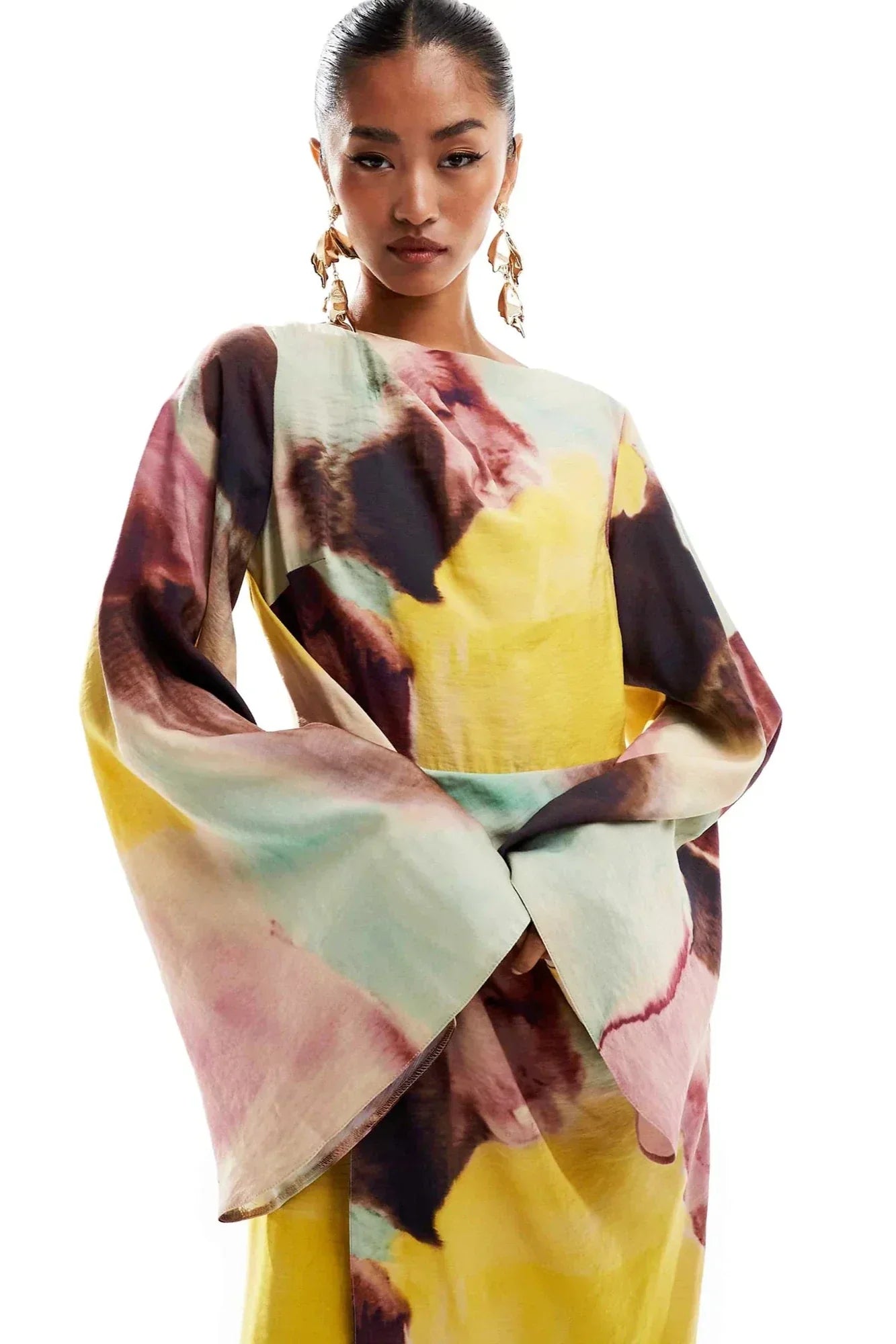 Watercolor - A woman models a long-sleeved maxi dress featuring a high neckline and a vibrant abstract watercolour print in yellow, pink, and brown. The dress has a twist-front detail at the waist and dramatic flared sleeves.