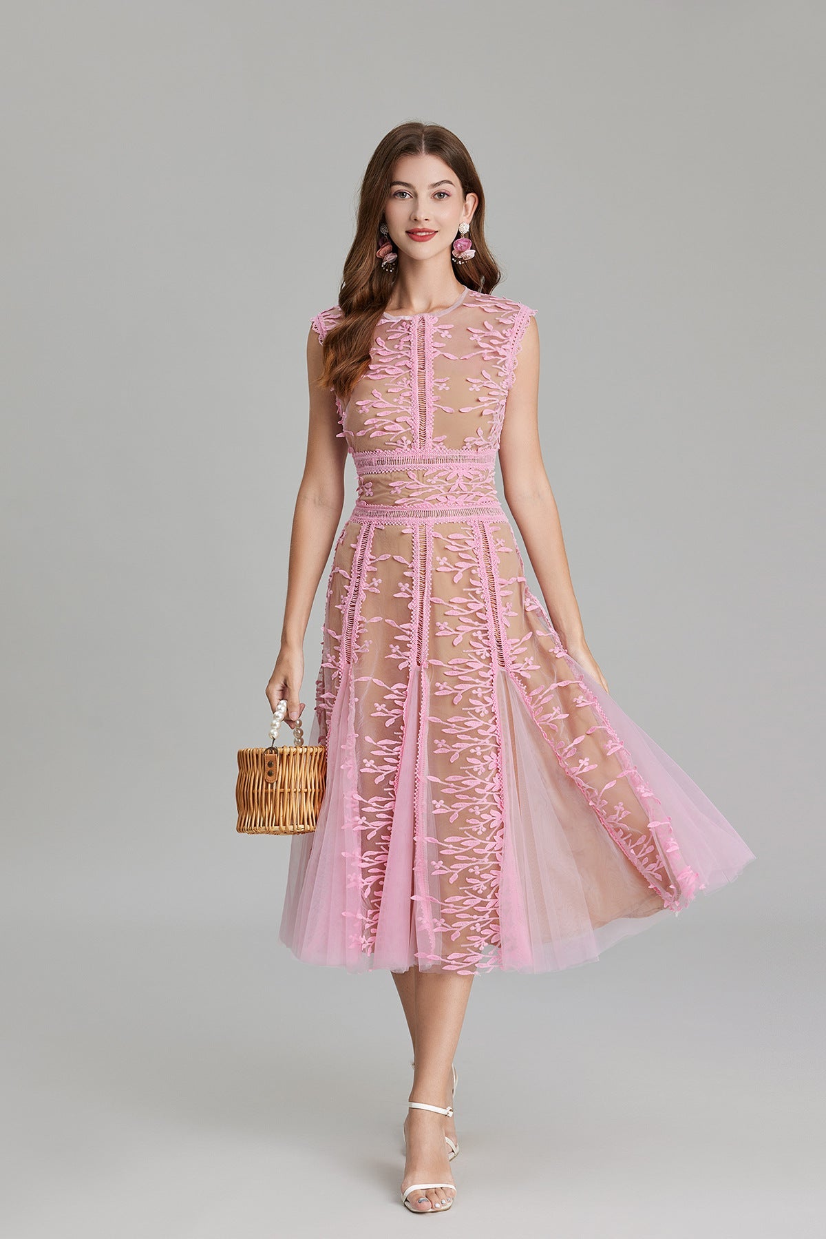 Full-length view of a woman wearing a sleeveless, sheer midi dress in pink and nude, featuring elaborate pink botanical embroidery in vertical panels and a flared tulle skirt.
