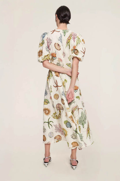 A woman models a short-sleeve, crewneck midi dress with a white background and a colorful, illustrative print of marine life including jellyfish, coral, and seashells.