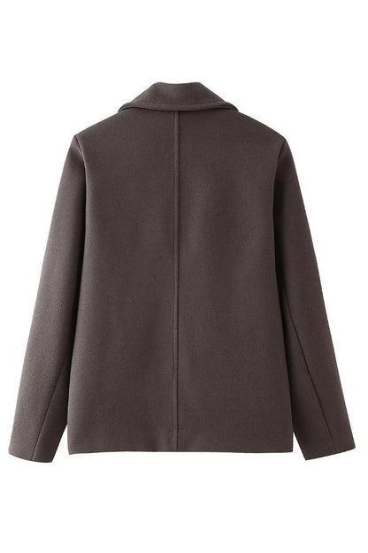 Grey - A woman wearing a taupe, short boxy jacket with oversized notch lapels, wide sleeves, and a double-breasted button front, styled over a black top.