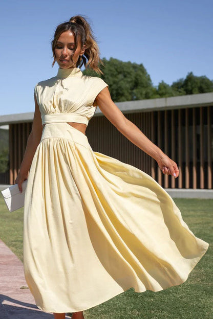 A woman walks outdoors in a pale yellow midi dress, its full skirt flowing around her. The dress features a high funnel neck, cap sleeves, and cutouts at the waist.