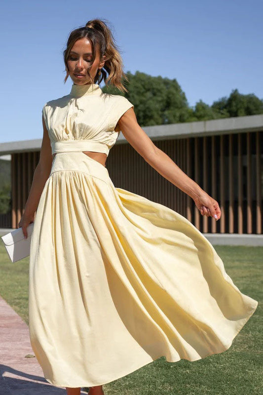 A woman walks outdoors in a pale yellow midi dress, its full skirt flowing around her. The dress features a high funnel neck, cap sleeves, and cutouts at the waist.