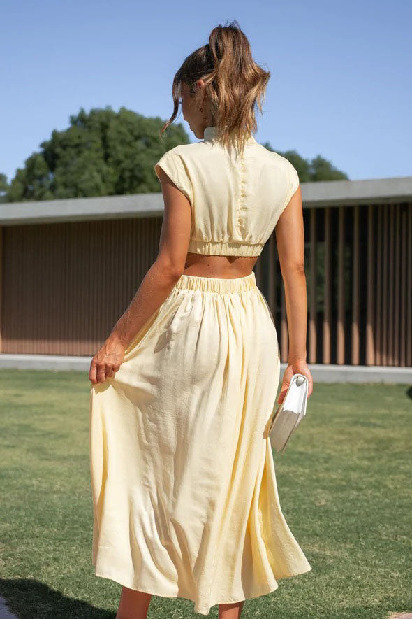A woman walks outdoors in a pale yellow midi dress, its full skirt flowing around her. The dress features a high funnel neck, cap sleeves, and cutouts at the waist.