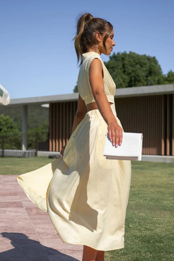 A woman walks outdoors in a pale yellow midi dress, its full skirt flowing around her. The dress features a high funnel neck, cap sleeves, and cutouts at the waist.