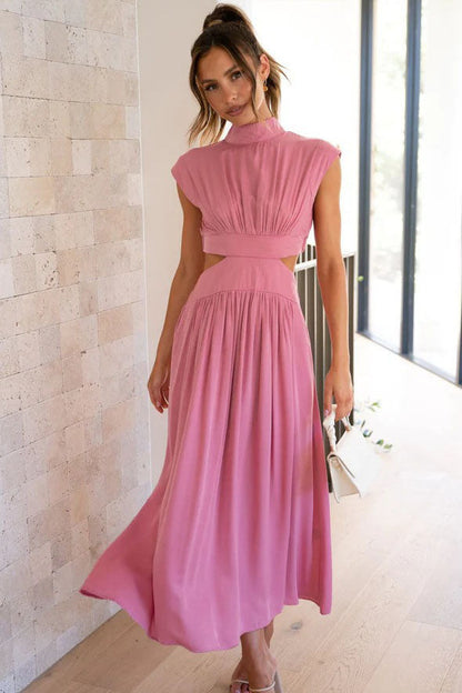 A woman walks outdoors in a pale yellow midi dress, its full skirt flowing around her. The dress features a high funnel neck, cap sleeves, and cutouts at the waist.