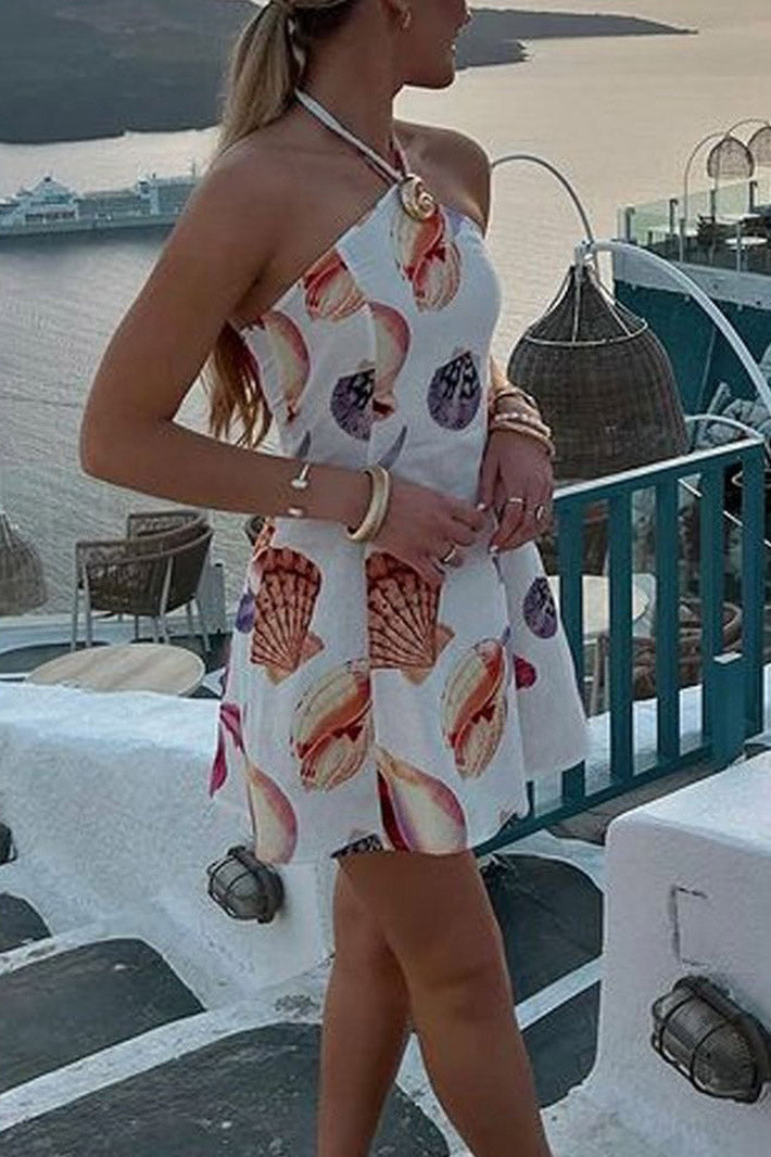 A woman wearing a white seashell print halter mini dress with a large gold spiral embellishment at the neckline, standing on a balcony overlooking the sea at sunset.