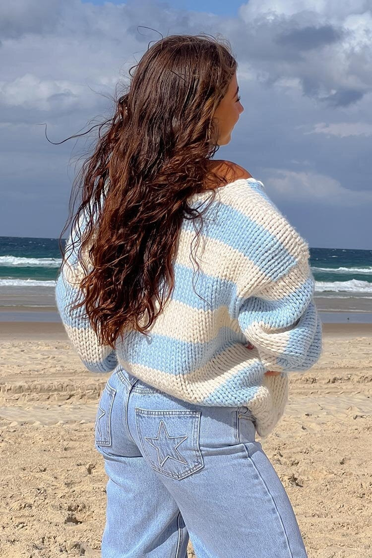 Blue - Woman wearing a chunky knit, slouchy off-shoulder jumper featuring wide horizontal stripes, paired with light wash denim jeans.