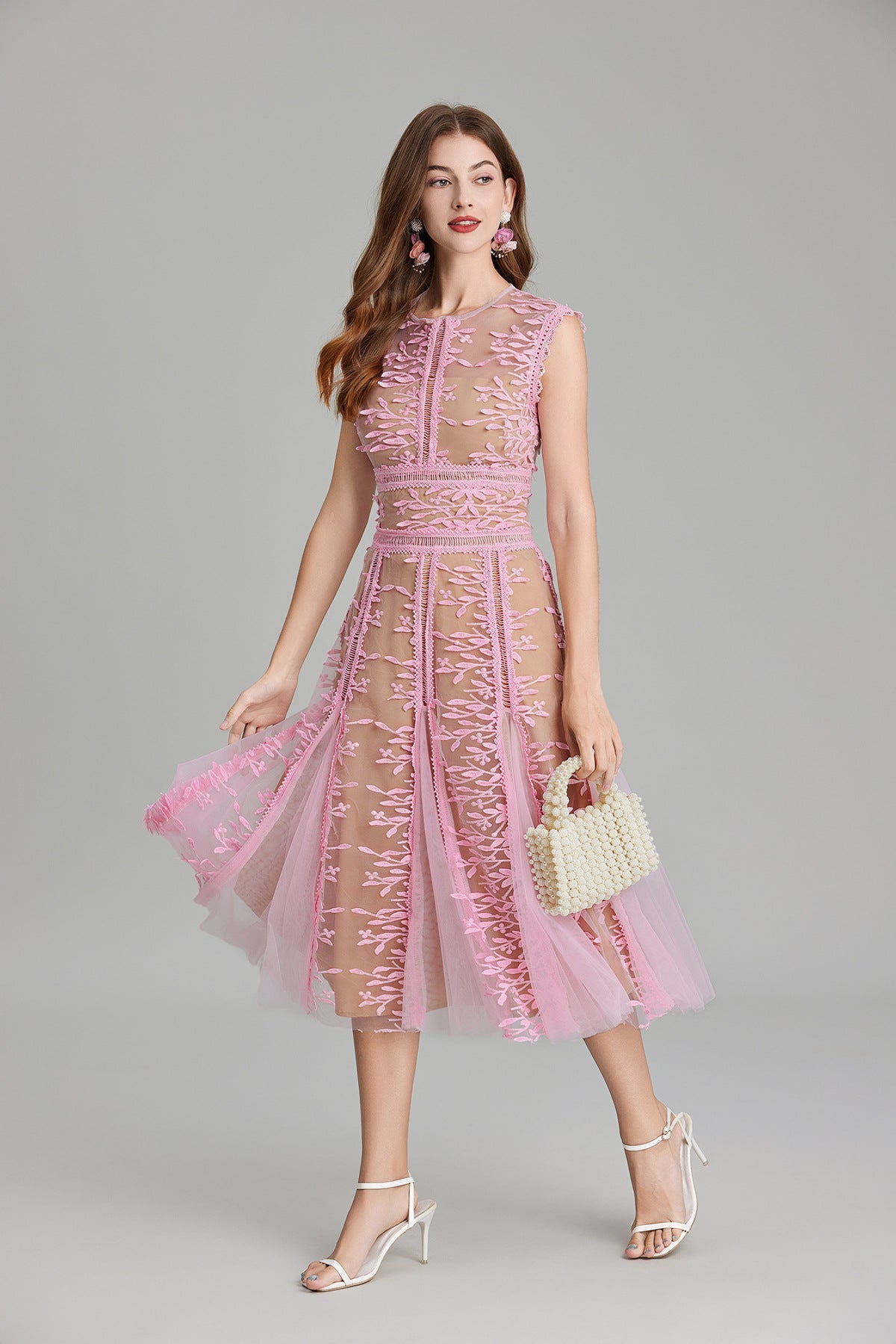 Full-length view of a woman wearing a sleeveless, sheer midi dress in pink and nude, featuring elaborate pink botanical embroidery in vertical panels and a flared tulle skirt.