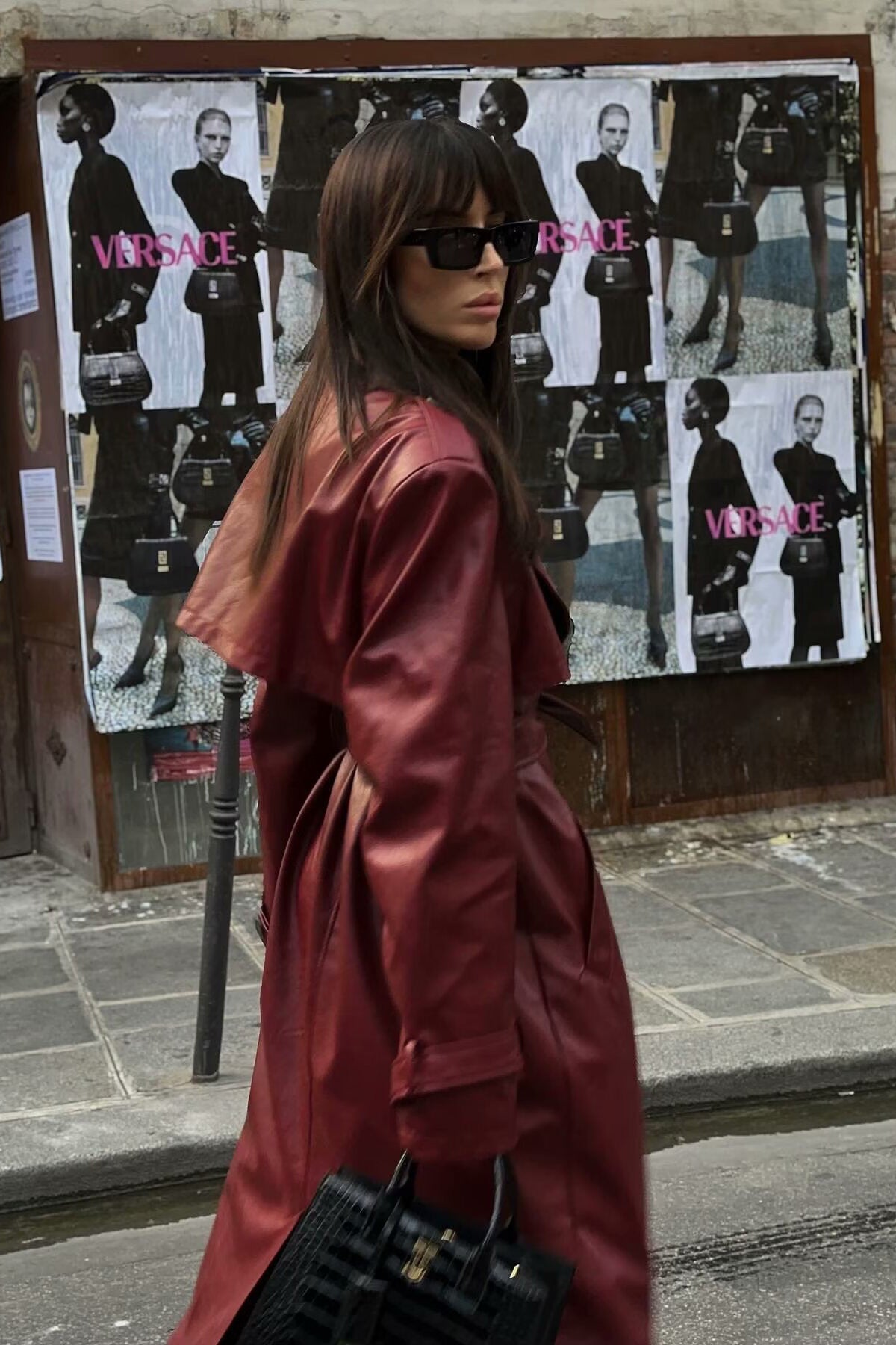 A woman walking down a city street wearing a long, deep burgundy faux leather trench coat with a tie belt, styled over a black turtleneck and black leggings.