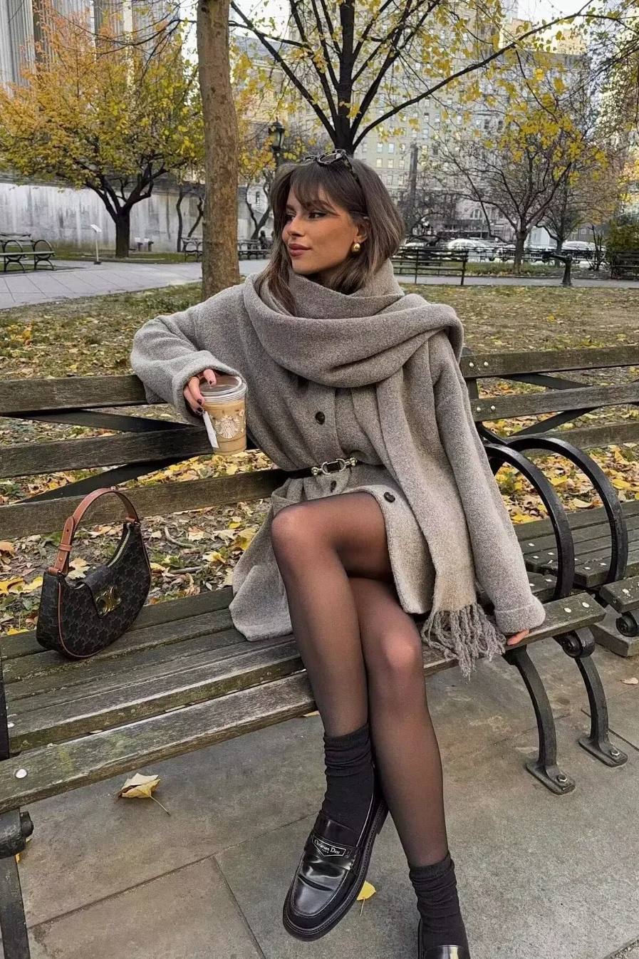 Khaki - A woman wearing a thick, taupe oversized longline winter coat featuring a built-in scarf neckline, dark button closures, and large patch pockets.