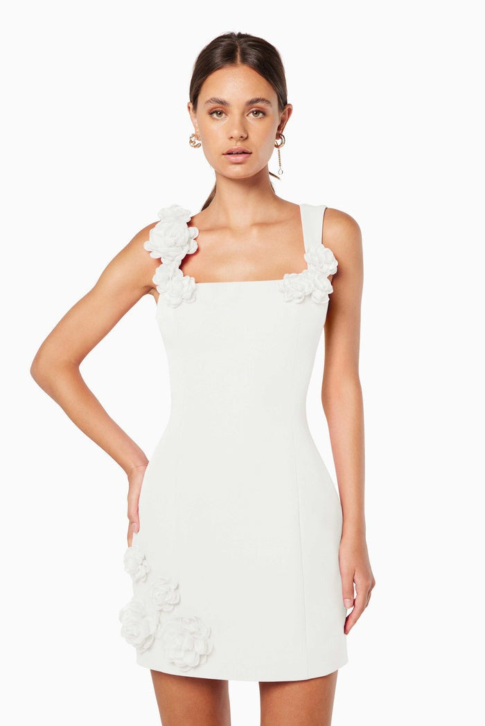 A woman models a white sleeveless A-line mini dress with a square neckline against a white background. The dress is detailed with white 3D floral appliqués on the straps and the left hip.
