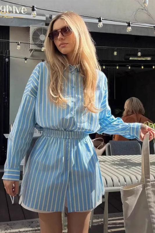A woman wearing a blue and pale yellow vertically striped mini shirt dress featuring a collared neckline, button-down front, long sleeves, and a thick elasticated waistband.