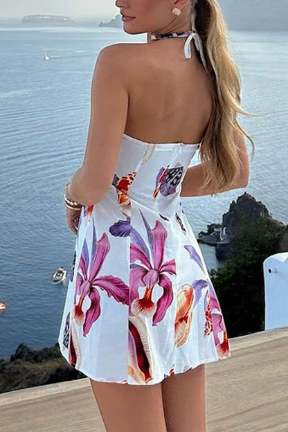 A woman wearing a white seashell print halter mini dress with a large gold spiral embellishment at the neckline, standing on a balcony overlooking the sea at sunset.