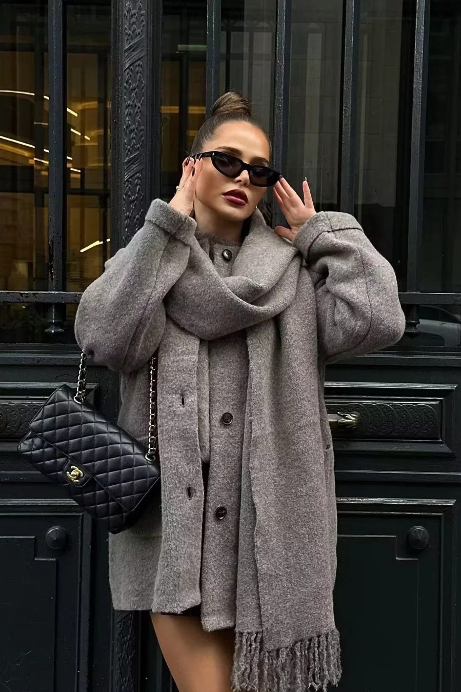 Grey - A woman wearing a thick, taupe oversized longline winter coat featuring a built-in scarf neckline, dark button closures, and large patch pockets.