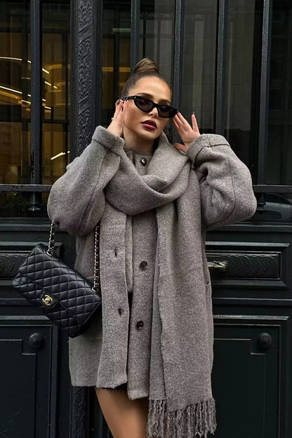 Grey - A woman wearing a thick, taupe oversized longline winter coat featuring a built-in scarf neckline, dark button closures, and large patch pockets.