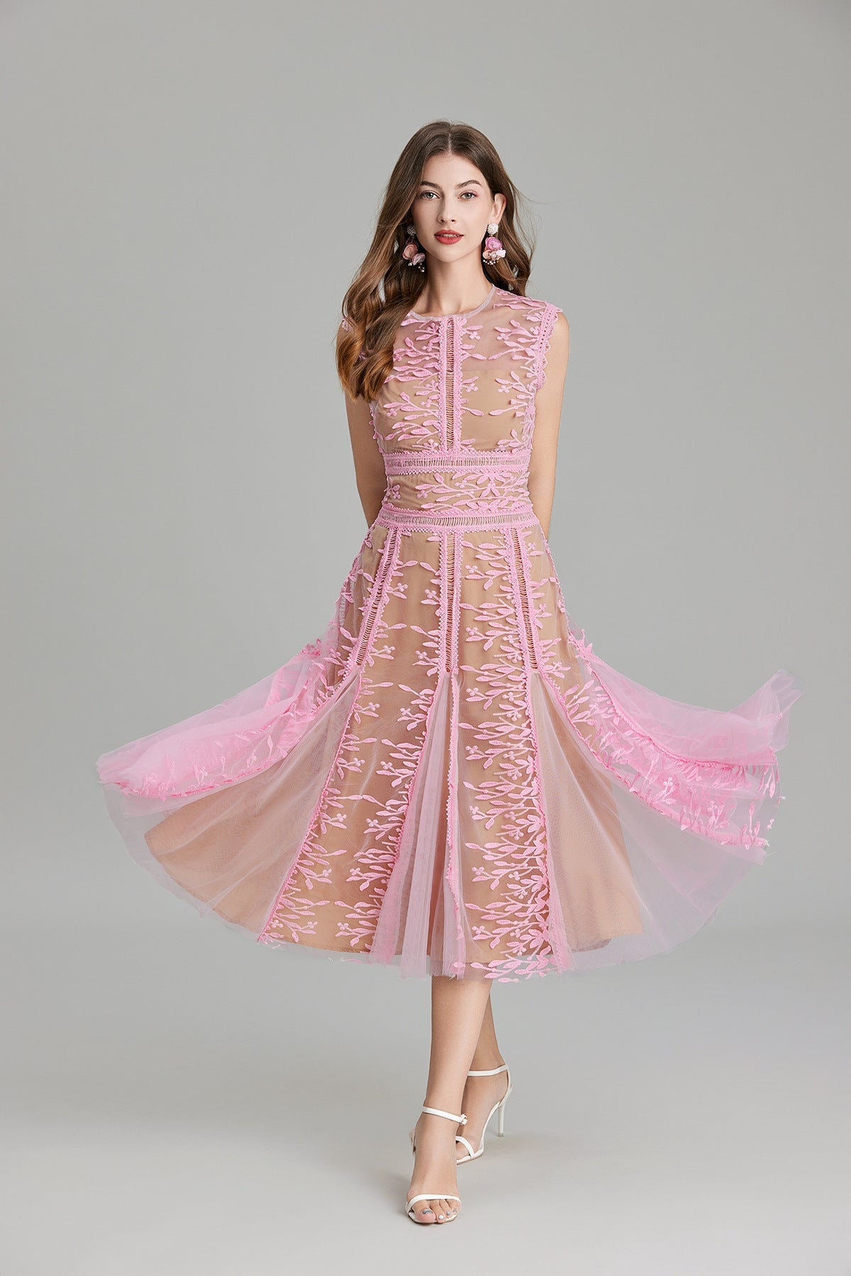Full-length view of a woman wearing a sleeveless, sheer midi dress in pink and nude, featuring elaborate pink botanical embroidery in vertical panels and a flared tulle skirt.