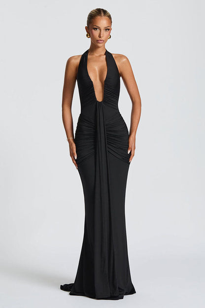 Black - A woman models a form-fitting, deep red halterneck maxi dress with a plunging neckline and extensive ruching, standing elegantly in a formal garden setting.