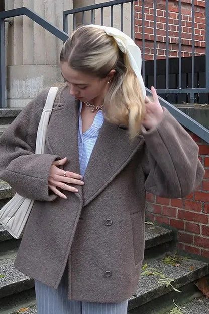 Grey - A woman wearing a taupe, short boxy jacket with oversized notch lapels, wide sleeves, and a double-breasted button front, styled over a black top.