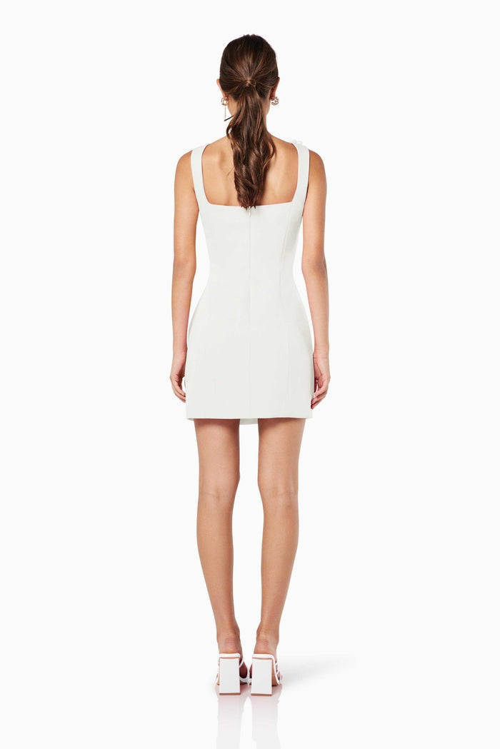 A woman models a white sleeveless A-line mini dress with a square neckline against a white background. The dress is detailed with white 3D floral appliqués on the straps and the left hip.