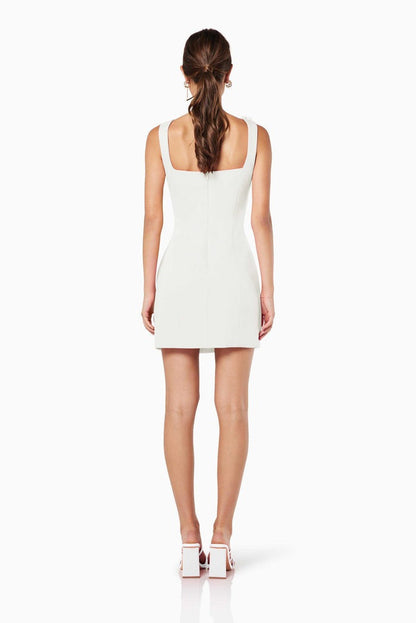 A woman models a white sleeveless A-line mini dress with a square neckline against a white background. The dress is detailed with white 3D floral appliqués on the straps and the left hip.