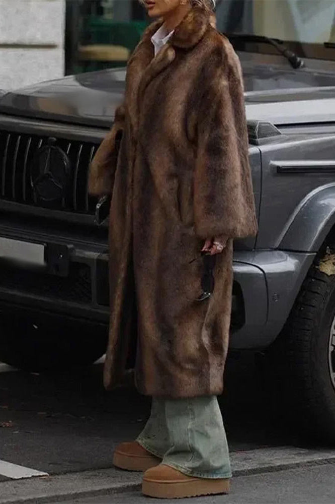 A woman wearing a long, oversized brown faux fur coat with a wide notch lapel, styled over a white shirt and jeans, standing in front of a dark SUV.